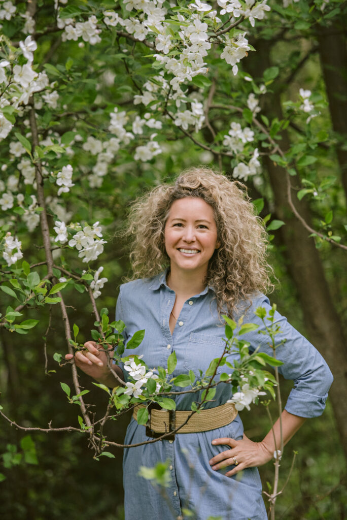 Smiling woman with curly hair stands among blossoming trees.