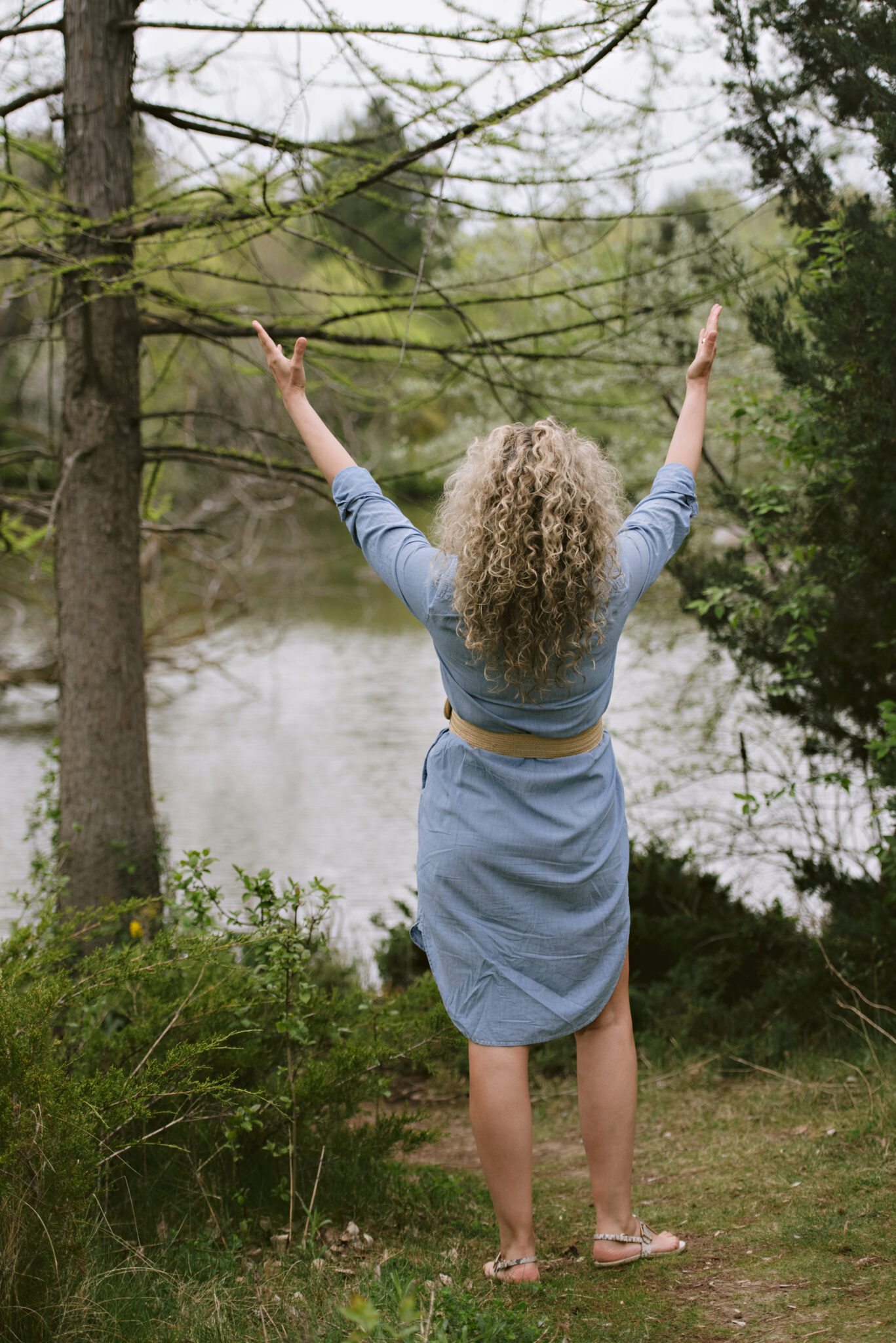 Woman with curly hair raising arms near a calm lake in nature.