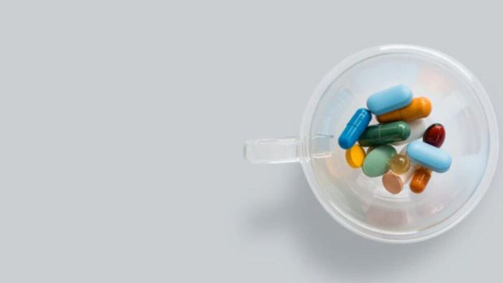 Various colorful capsules and pills in a transparent cup on a gray background.