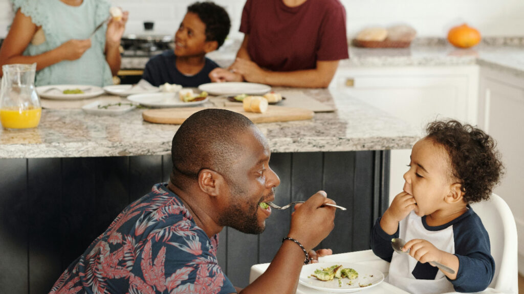 A man joyfully feeds a child at the dining table.